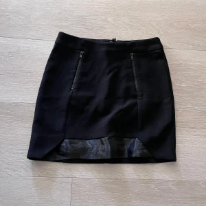 Svart kortkjol med dragkedjor - Snygg svart kortkjol med två dragkedjor framtill och en liten slits med glansigt tygdetalj längst ner. Perfekt att styla med en cool topp eller stickad tröja.