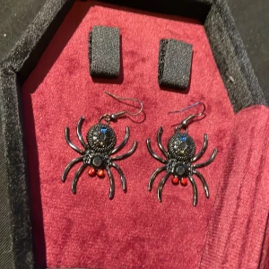 Svarta spindelörhängen med röda detaljer - Coola örhängen formade som svarta spindlar med små röda detaljer. Perfekta för dig som gillar unika accessoarer med lite edge. Kommer i en snygg kistformad ask med röd sammet.