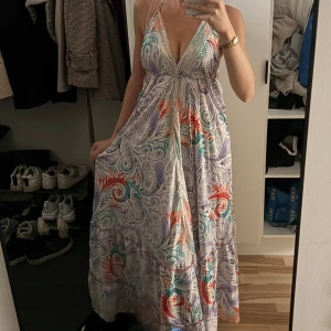 Långklänning med paisleymönster i pastellfärger - Säljer en flowy långklänning med djup v-ringning och halterneck. Klänningen har ett färgglatt paisleymönster i lila, turkos, röd och orange på ljus botten. Perfekt för sommarens alla tillfällen!