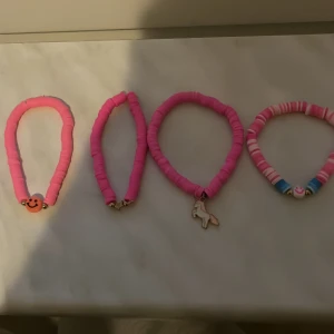 Fyra rosa armband med olika berlocker - Säljer fyra söta armband i olika nyanser av rosa. Två av armbanden har berlocker, en med en glad smiley och en med en enhörning. Ett armband har färgglada detaljer i blått och vitt. Perfekta accessoarer för att liva upp din outfit!