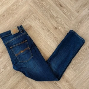 Blå jeansbyxor från Nudie Jeans - Säljer ett par klassiska blå jeans från Nudie Jeans med fem fickor och normal passform. Jeansen har en snygg tvätt och subtila slitningar, samt Nudie-logga på bakfickan. Perfekta för en avslappnad stil.