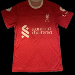 Liverpool FC matchtröja M. Salah 11 - Röd Liverpool FC fotbollströja med vita detaljer, tryck av M. Salah och nummer 11 på ryggen. Kortärmad modell med klubbmärke och sponsorlogga på bröstet. Klassisk rund hals och diskret mönster i tyget.