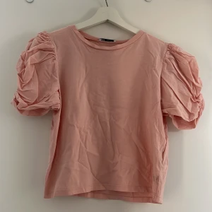 Rosa topp med puffärmar från ZARA - Säljer en ljusrosa topp från ZARA med korta, voluminösa puffärmar. Toppen har en rak passform och är tillverkad i mjuk bomull. Inga defekter och använd några fåtal gånger🩷