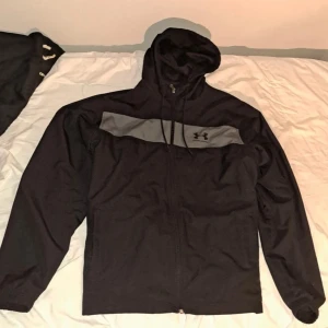 Svart vindjacka med huva från Under Armour - Säljer en svart vindjacka från Under Armour med grått bröstparti och logga. Jackan har huva med dragsko och dragkedja framtill. Perfekt för träning eller när det blåser ute.