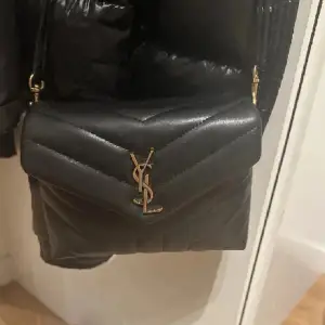 Säljer en svart axelväska från Saint Laurent med quiltad design och guldigt YSL-spänne framtill. Väskan har en elegant, klassisk form och justerbar axelrem. Perfekt för att lyfta vilken outfit som helst.
