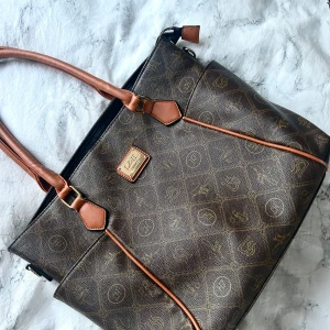 Brun handväska  - Säljer en brun handväska med mönstrad yta och ljusbruna handtag. Väskan har dekorativa detaljer i guld och praktiska fack. Perfekt för dig som vill ha en stilren accessoar med klassisk känsla. 