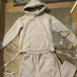 Carsicko tracksuit  - Säljer ett par ljusgrå mjukisbyxor från Cenario med resår i midjan och ett broderat hjärta med ansikte på ena benet. Byxorna har avslappnad passform och är perfekta för en chill stil.