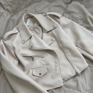 Beige skinnjacka med dragkedjor - Snygg beige skinnjacka i bikerstil med silvriga dragkedjor och knappar. Jackan har en klassisk krage, lång ärm och detaljerade sömmar. Perfekt för att lyfta vilken outfit som helst.