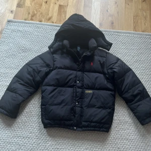Svart dunjacka med huva från Polo Ralph Lauren - Svart pufferjacka från Polo Ralph Lauren med huva och röd broderad logga på bröstet. Jackan har dragkedja och knappar. Perfekt för kyliga dagar.