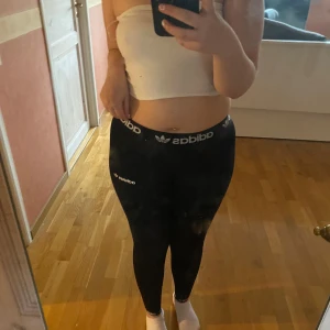 Svarta leggings från Adidas - Säljer ett par svarta leggings från Adidas med vit logga och text på midjan. De har en tight passform och är perfekta för träning eller en sportig look. Klassisk design med hög midja och stretchigt material.