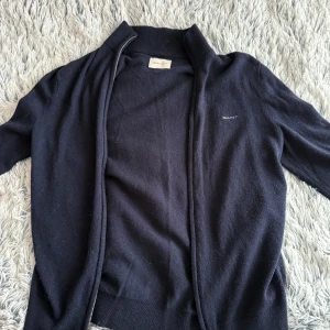 Mörkblå zip kofta från GANT - Säljer en klassisk mörkblå zip kofta från GANT i storlek S. Koftan har dragkedja framtill, ribbade muddar och hög krage. Perfekt för lager-på-lager och enkel att matcha med olika outfits.