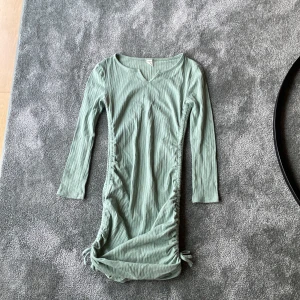 Ljusgrön ribbad klänning från SHEIN - Säljer en ljusgrön ribbad klänning från SHEIN med lång ärm och v-ringning. Klänningen har dragsko och knyt i sidorna nertill för en snygg detalj. Perfekt för dig som gillar en enkel men trendig stil.
