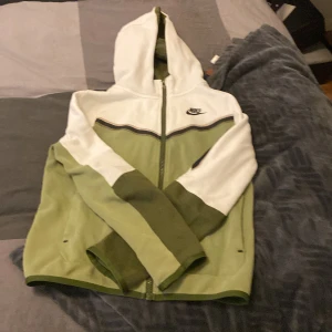 Nike hoodie med dragkedja i grönt och vitt - Säljer en snygg hoodie från Nike i grönt och vitt med dragkedja framtill och huva. Jackan har svarta detaljer, ficka på ärmen och klassisk Nike-logga på bröstet. Perfekt för en sportig stil.