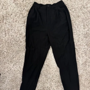 Svarta kostymbyxor med knapp - Säljer ett par svarta kostymbyxor med hög midja och klassisk passform. Byxorna har hällor för bälte och stängs med knapp och dragkedja framtill. Perfekta för en stilren och enkel look.