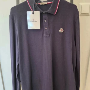 Mörkblå långärmad pikétröja från Moncler - Säljer en mörkblå långärmad pikétröja från Moncler med klassisk krage och knappar framtill. Kragen har röda och vita detaljer och Moncler-loggan sitter på bröstet. Perfekt för en stilren och sportig look.