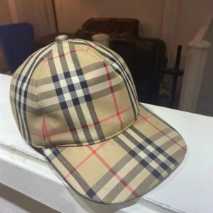 Beige rutig keps från Burberry - Snygg keps från Burberry med klassiskt beige rutigt mönster och justerbar baksida. Märkt med 'Burberry London England' baktill. Perfekt accessoar för en trendig look.