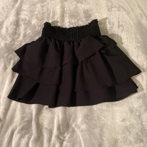 Svart volangkjol med resårmidja - Supersöt svart kjol med flera lager volanger och bred smockresår i midjan. Kjolen har en luftig känsla och passar perfekt till både vardag och när du vill känna dig extra fin. Enkel att matcha med olika toppar.