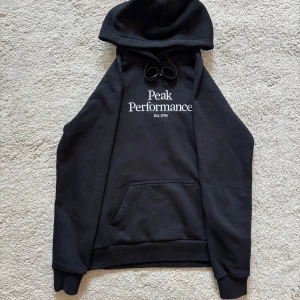 Svart hoodie från Peak Performance - Svart hoodie från Peak Performance med broderad vit logga på bröstet. Klassisk känguruficka, snörning i huvan och mjukt material. Perfekt för chill dagar eller när du vill ha en clean och stilren look.