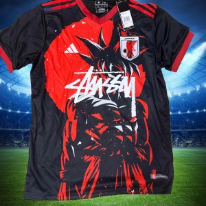 Japan x Stüssy x Adidas fotbollströja  - Unik Japan fotbollströja i samarbete med Stüssy och Adidas. Svart bas med röd och vit grafisk design framtill, inspirerad av anime och streetwear. Korta ärmar, röd krage och ärmslut. Tillverkad i lätt och ventilerande material (AEROREADY).
