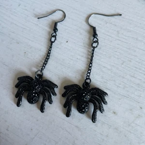 Svarta örhängen med spindelmotiv - Coola svarta örhängen med hängande spindlar i glittrigt material. Kedjorna ger extra edge och spindlarna har en tydlig, detaljerad form. Perfekta för dig som gillar unika accessoarer med lite attityd.