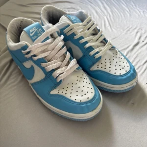 Nike SB Dunk Low Pro blå/vit sneakers - Nike SB Dunk Low Pro sneakers i blått och vitt med klassisk låg siluett. Skorna har vit bas med blå overlays, perforerad tåbox och vit snörning. Yttermaterial i skinn och slitstark sula. Perfekta för dig som gillar streetstyle och skate.