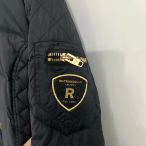 Svart lång dunjacka Rockandblue - Lång svart dunjacka från Rockandblue med quiltade partier och guldiga dragkedjor. Jackan har huva, flera fickor med dragkedja och en snygg patch på ärmen. Perfekt för kalla dagar när du vill hålla stilen och värmen.