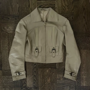 Beige croppad skinnjacka med bälten - Snygg retro beige croppad skinnjacka med spännen. Jackan är i äkta läder! Den har små diskreta färgfläckar (man kan se dem om man zoomar in på ärmen på första bilden). Skriv för fler bilder (tex närmare på fläckarna). Jackan är köpt secondhand av min mamma när hon var ung och den är fortfarande hel och fin bortsett från de små fläckarna!