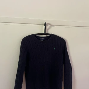 Mörkblå stickad tröja Polo Ralph Lauren - Kabelstickad mörkblå tröja från Polo Ralph Lauren med rund halsringning och lång ärm. Klassisk design med grönt broderat logomärke på bröstet. Perfekt för dig som gillar stilrena och tidlösa plagg.