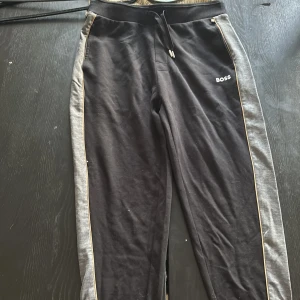 Svarta mjukisbyxor från BOSS - Svarta mjukisbyxor från BOSS med gråa sidor och tunna guldiga detaljer längs benen. Byxorna har snörning i midjan och BOSS-logga på vänster höft. Perfekta för en chill och sportig look.