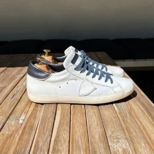 Vita sneakers Philippe Model - Snygga vita sneakers från Philippe Model med mörkblå detaljer på häl och tunga. Skorna har blå snörning, rund tå och platt sula. Tillverkade i skinn för en clean och stilren look, perfekta till jeans eller shorts. Nypris: 3400kr• Skick: 9/10