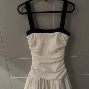 Vit och svart klänning från Zara - Supersnygg kort klänning från Zara i vit med breda svarta axelband och kant upptill. Klänningen har en figurnära passform upptill och en lätt volangig kjol. Perfekt för dig som vill sticka ut med en stilren look. Köp innan fredag så postar jag tills dess. 
