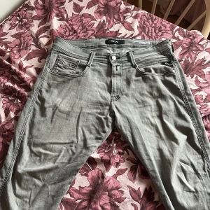 Replay Anbass grå slim jeans - Snygga grå jeans från Replay, modell Anbass med slim fit. Klassisk femficksdesign, diskreta sömmar och logga på bakfickan. Jeansen är i mjuk bomullsdenim och passar perfekt till en avslappnad streetstil. Dom är omsydda i benen för att dom var för långa så man kan säga att storleken i längden är ca 31.