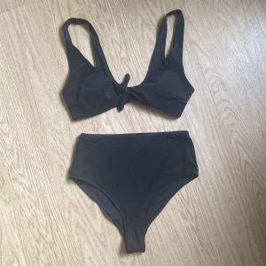 Svart ribbad bikini från Kay/Day (Kappahl) storlek XS - Snygg svart bikini från Kay/Day (Kappahl) i storlek XS. Överdelen har breda axelband och en knytning framtill, medan underdelen är high waist. Materialet är ribbat och stretchigt för en bekväm passform. Perfekt för strandhäng eller poolparty!