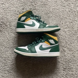 Nike Air Jordan 1 Mid grön/vit/gul - Nike Air Jordan 1 Mid sneakers i grönt, vitt och gult. Klassisk high-top siluett med gröna läderpaneler, vit bas och gula detaljer vid ankeln. Ikonisk Jordan-logga på sidan och gröna snören. Perfekta för dig som vill sticka ut med en retro basketstil.