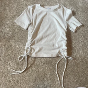 Vit ribbad topp med snörning från Zara - Superfin vit ribbad topp från Zara med korta ärmar och snygg snörning i sidorna som kan justeras för en cool look. Mjuk och stretchig, perfekt till jeans eller kjol. Enkel men ändå trendig design som passar året runt.