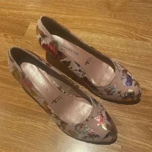 Blommiga pumps från Tamaris, stl 39 - Snygga pumps från Tamaris i beige med metallic blommönster i guld, rött, lila och grönt. Skorna har rund tå, låg klack och är tillverkade i ett glansigt syntetmaterial. Perfekta för dig som vill sticka ut med en färgglad och trendig look. Tryck ej köp nu
