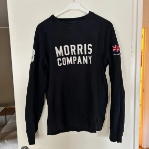 Svart sweatshirt från Morris Company - Svart sweatshirt från Morris Company med vit broderad text framtill och brittisk flagga på ärmen. Klassisk rund halsringning och långa ärmar. Tillverkad i mjuk bomull, perfekt för en chill och stilren look. Kostade 1299kr ny