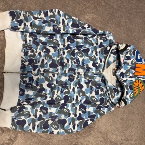 BAPE camo zip hoodie shark S - Säljer en ikonisk BAPE zip hoodie i blå camo med hajmotiv på huvan och broderade bokstäver. Jackan har hel dragkedja, kängurufickor och är tillverkad i mjuk bomull. Perfekt för dig som vill sticka ut med streetwear-vibe och är även perfekt för sommaren.