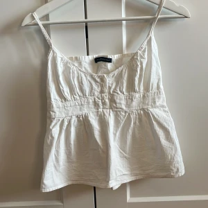 Vit linnetopp från Brandy Melville - Supersöt vit linnetopp från Brandy Melville med smala axelband och knappar framtill. Toppen har en lätt rynkad midja och luftig passform, perfekt för varma dagar. Tillverkad i mjuk bomull med diskret struktur i tyget.
