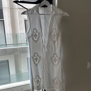 Vit ärmlös klänning från Zara oanvänd  - Vit ärmlös kortklänning från Zara med broderade detaljer i beige och snörning framtill. Klänningen har krage och ett luftigt material som passar perfekt för varma dagar. Snygg och stilren med unika broderier som ger en bohemisk vibe. 