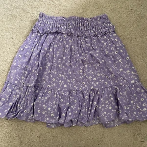 Ljuslila blommig volangkjol från Zara - Supersöt ljuslila kortkjol med vita små blommor och volangkant från Zara. Kjolen har en bred, smockad midja som ger en bekväm och snygg passform. Perfekt för vår och sommar när du vill känna dig extra fin.