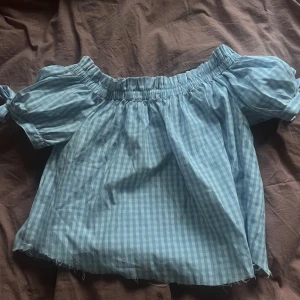Blå rutig offshoulder blus - Supersöt blå och vit rutig blus med offshoulder och puffiga korta ärmar. Toppen har resår vid axlarna och en luftig, croppad passform. Perfekt för varma dagar och ger en riktigt somrig vibe.