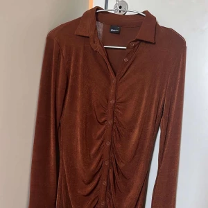 Brun sammetsklänning från Gina Tricot - Snygg brun sammetsklänning med långa ärmar och krage från Gina Tricot. Klänningen har knappar hela vägen framtill och cool rynkdetalj längs mitten som ger en unik look. Perfekt för dig som vill sticka ut lite extra.