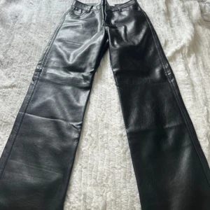 Svarta skinnbyxor från Bershka - Snygga svarta skinnbyxor från Bershka i rak modell. Byxorna har hög midja, klassiska fickor och bälteshällor. Materialet är syntetiskt skinn som ger en cool look. Perfekta för dig som vill sticka ut med en edgy vibe. aldrig använda helt nya 