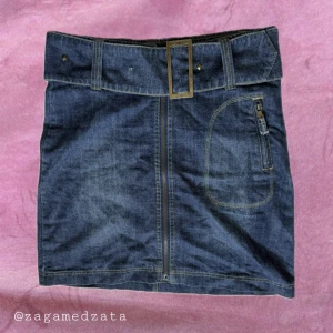 Jeanskjol med bälte från SWE Street - Mörkblå jeanskjol från SWE Street med bredt bälte och metallspänne. Kjolen har framficka med dragkedja och två bakfickor med nitar. Klassisk denimlook som är lätt att styla till olika outfits.