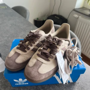 Adidas Samba  - Adidas Samba sneakers i snygg beige och brun färgkombination. Ovandel i mjuk mocka med klassiska tre ränder i brunt och matchande bruna skosnören. Guldtexten 'SAMBA' på sidan ger en extra touch. Platt sula och rund tå för en clean look.
