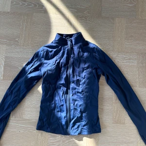 Mörkblå tight sportjacka med dragkedja - Snygg mörkblå vindjacka med hel dragkedja framtill och hög krage. Jackan har en slim passform och långa ärmar, perfekt för blåsiga dagar. Enkel och stilren design som funkar till många outfits.