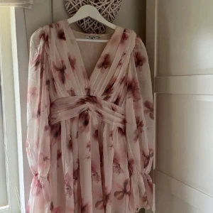 Klänning från NA-KD - Superfin kortklänning från NA-KD i ljusrosa med blommigt mönster i vinröda toner. Klänningen har v-ringning, markerad midja och långa, transparenta ärmar. Perfekt till midsommar och endast andävnd 1 gång!!❤️Nypris 700 kr🥰