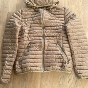 Beige pufferjacka från Colmar - Snygg beige pufferjacka från Colmar med horisontella sömmar, huva och dragkedja framtill. Jackan har två dragkedjeförsedda fickor och en diskret logga på ärmen. Perfekt för kyliga dagar och enkel att matcha med olika outfits.