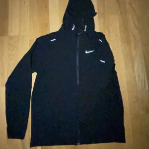 Svart Nike vindjacka med huva - Svart vindjacka från Nike med hel dragkedja och huva. Jackan har reflekterande detaljer på bröstet, axlarna och ärmsluten. Perfekt för löpning eller utomhusaktiviteter. Långärmad och sportig design med klassisk Nike-logga på bröstet.
